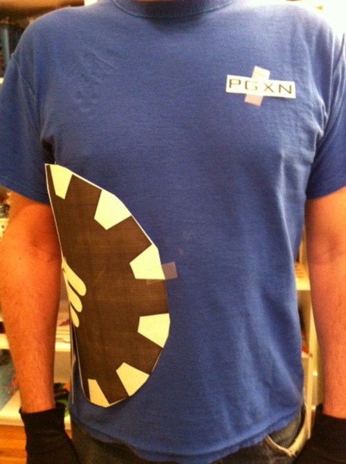 Photo of a male torso in a blue-ish shirt with the PGXN logo pinned to its side